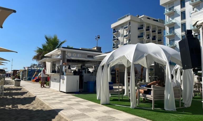 Spiaggia con bar, gazebo e palme sotto un cielo azzurro.