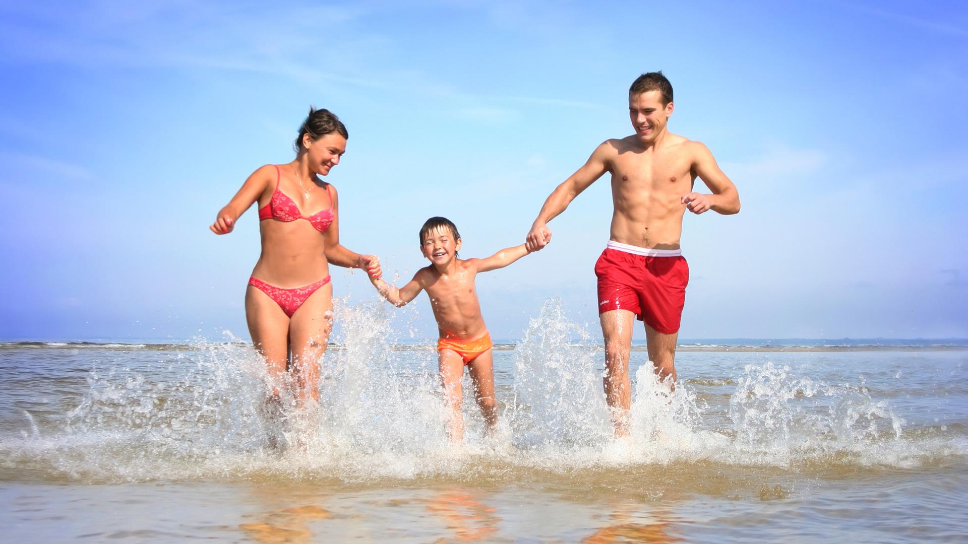 Famiglia felice corre in acqua al mare, cielo azzurro e onde leggere.